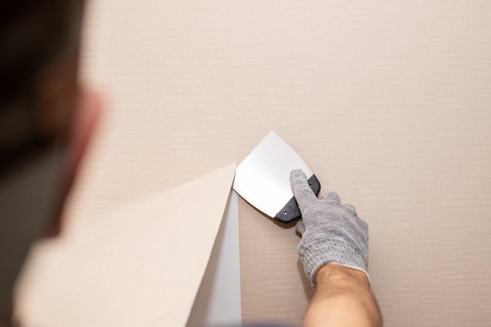Een persoon gebruikt een plamuurmes om glasvliesbehang voorzichtig van de muur te schrapen, terwijl de muur nat is van het water dat is aangebracht om het behang los te maken.