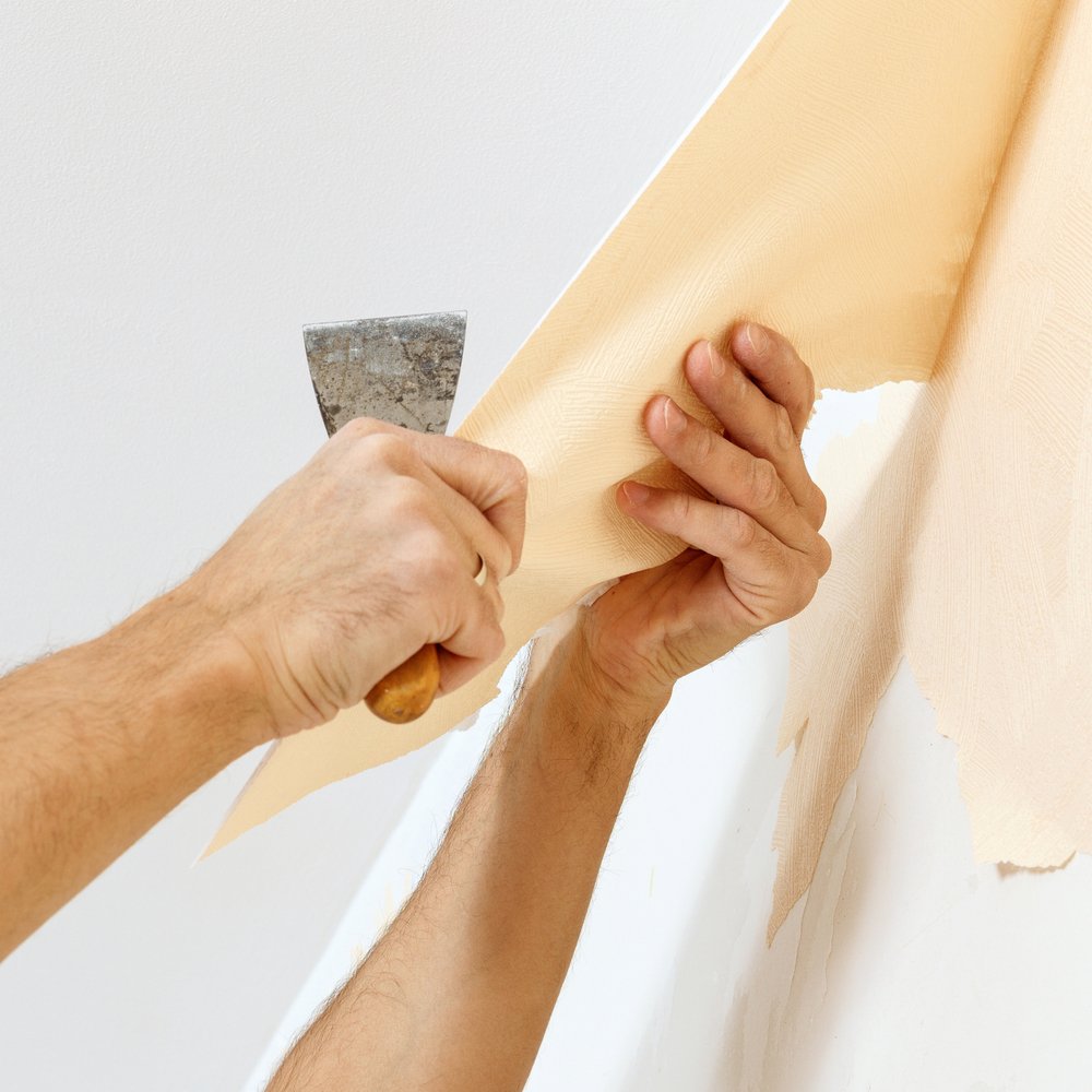 Een persoon gebruikt een plamuurmes om scanbehang voorzichtig van de muur te schrapen, terwijl de muur nat is van het water dat is aangebracht om het behang los te maken.