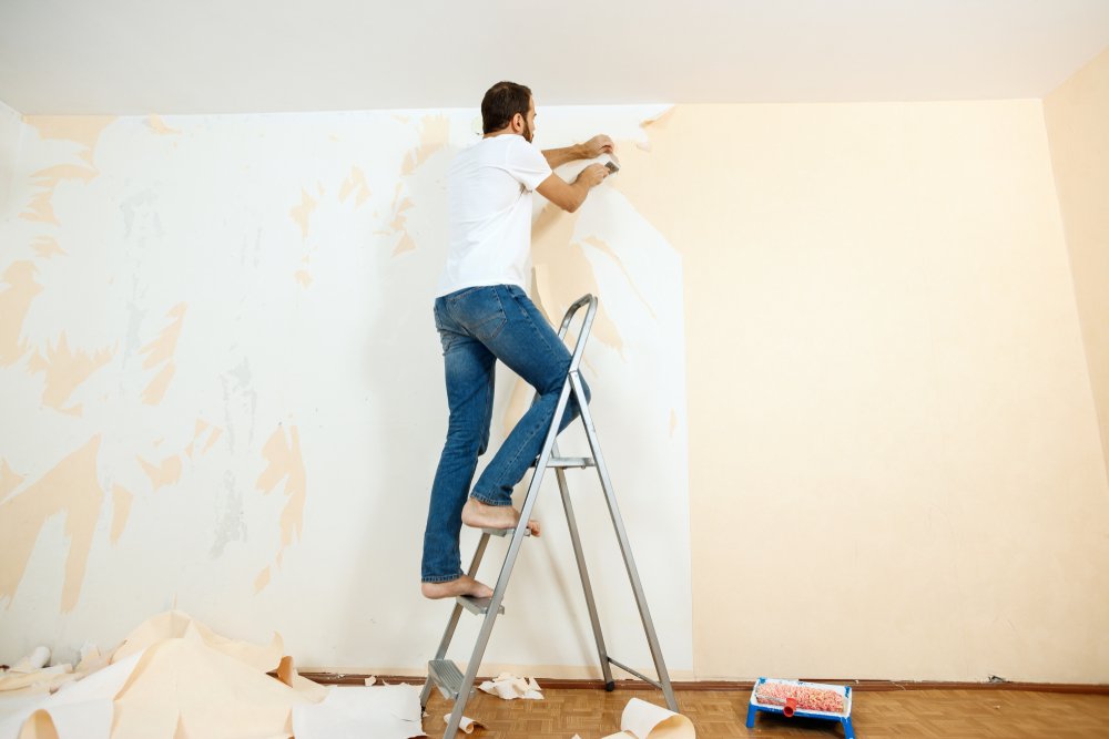 Een persoon gebruikt een plamuurmes om glasweefselbehang voorzichtig van de muur te schrapen, terwijl de muur nat is van het water dat is aangebracht om het behang los te maken.